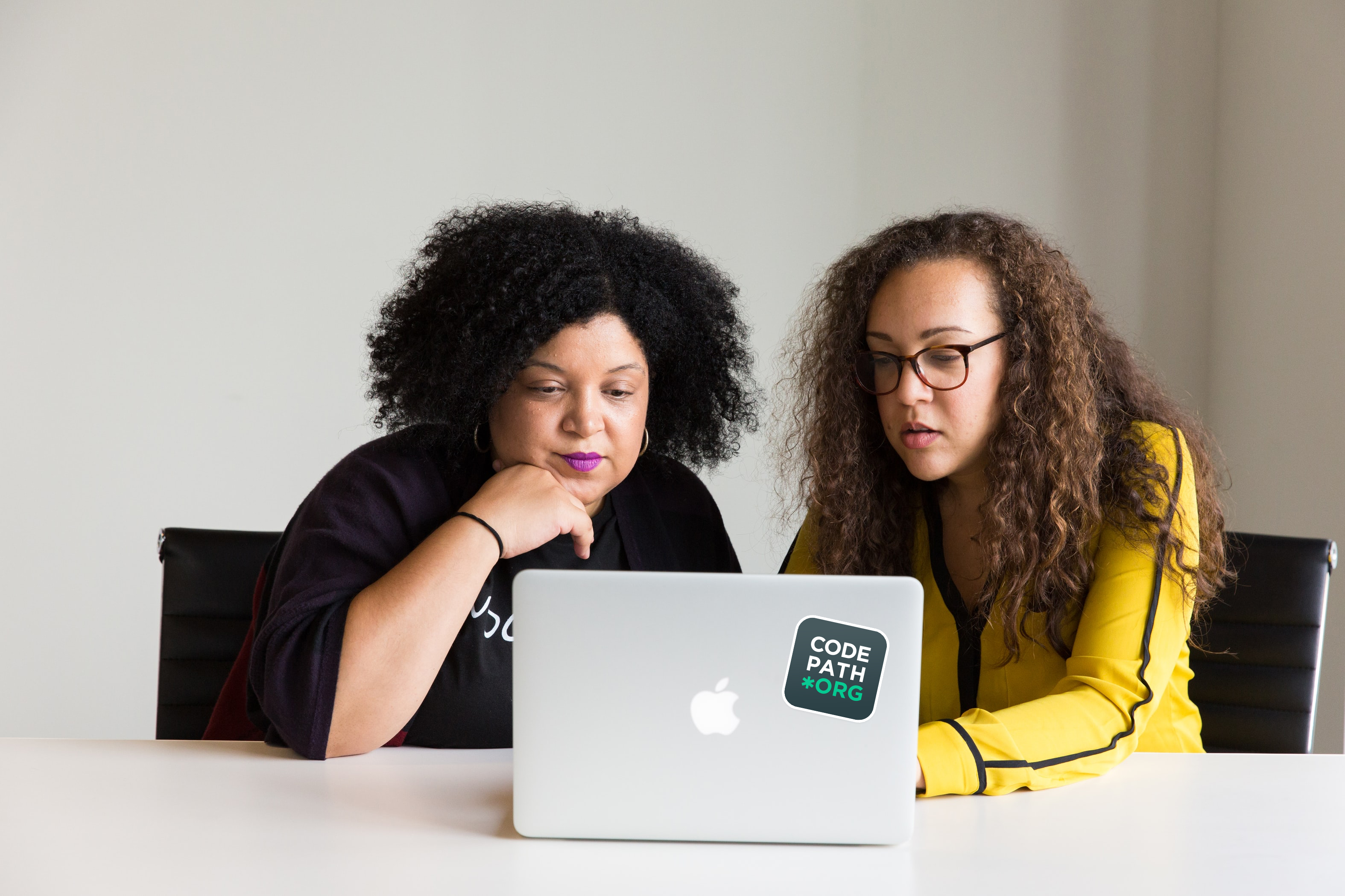 Two people sit in front of a laptop that has a CodePath sticker on the back. It is assumed these individuals are software engineers working collaboratively a project
